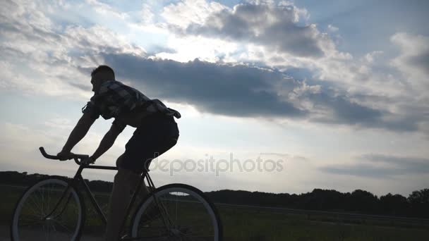 年轻男子骑在老式自行车与美丽的夕阳的天空背景下的剪影。在乡村的小路上骑自行车运动的家伙。踏单车的男子骑自行车。健康积极的生活方式慢动作 — 图库视频影像