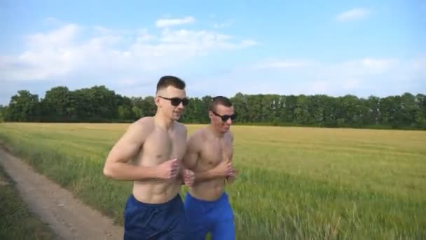 Dos hombres musculosos corriendo y hablando al aire libre. Jóvenes chicos atléticos sonrientes corriendo por el campo. Hombres deportistas entrenando juntos en la naturaleza. Amigos haciendo ejercicio afuera. Vida activa saludable — Vídeos de Stock