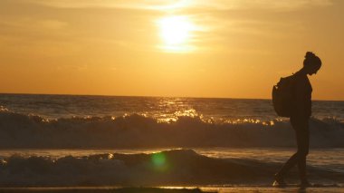 Genç kadın turist günbatımında Deniz Sahil boyunca yürüyüş sırt çantası ile. Güzel genç Gezgin kadın okyanus kıyısında güneş doğarken. Seyahat ya da yaz tatili sırasında atlama kız uzun yürüyüşe çıkan kimse