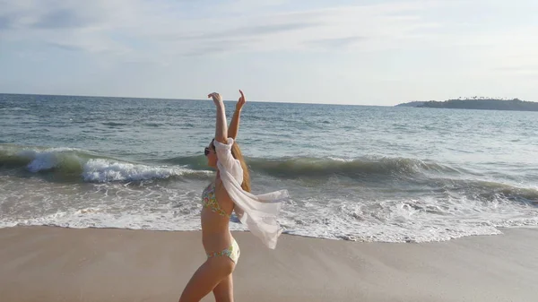Donna felice in bikini e camicia che cammina sulla spiaggia vicino all'oceano e alza la mano. Giovane bella ragazza godendo la vita e divertirsi sulla riva del mare. Vacanze estive o vacanze Paesaggio sullo sfondo — Foto Stock
