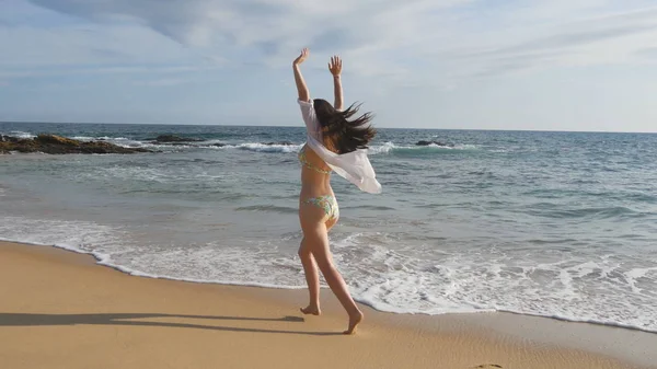 Donna felice in bikini e camicia che corre sulla spiaggia sabbiosa vicino all'oceano. Giovane bella ragazza godendo la vita e divertirsi sulla riva del mare. Vacanze estive o vacanze. Paesaggio naturale sullo sfondo — Foto Stock