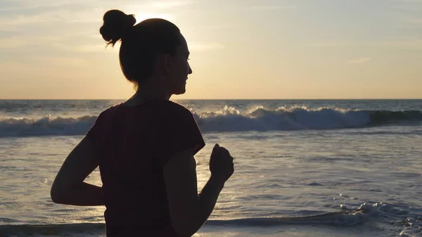 Silhouette de jeune femme courant sur la plage au coucher du soleil. Fille jogging le long de la côte de l'océan pendant le lever du soleil. Femme sportive faisant de l'exercice en plein air. Mode de vie sain et actif à la nature. Gros plan — Photo