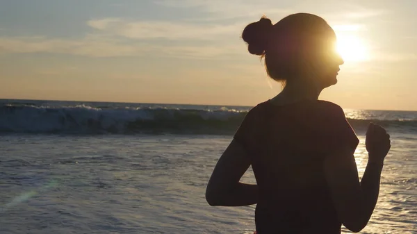 Silhouette di giovane donna che corre sulla spiaggia al tramonto. Ragazza che fa jogging lungo la riva dell'oceano durante l'alba. Sportivo femminile che si allena all'aperto. Stile di vita sano allenamento attivo alla natura. Da vicino. — Foto Stock