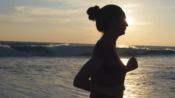 Sylwetka młoda kobieta działa na plaży o zachodzie słońca. Dziewczynka jogging wzdłuż brzegu oceanu podczas sunrise. Ćwiczeń odkryty kobiece sportowca. Styl życia zdrowy treningu o charakterze. Z bliska — Zdjęcie stockowe