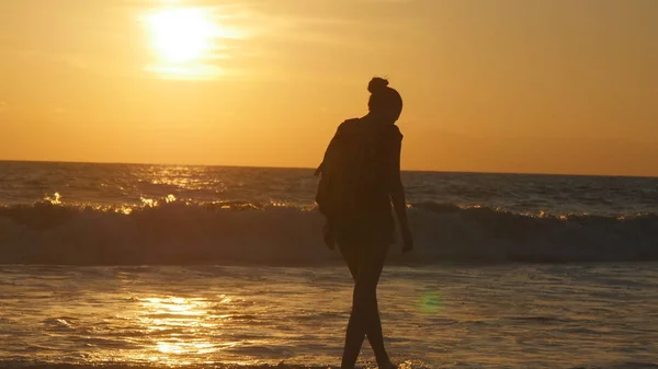 Ung kvinnlig turist med ryggsäck promenader längs stranden i solnedgången. Vackra unga resenären kvinna kommer på stranden i soluppgången. Flicka hiker kliva under resor eller sommar semester — Stockfoto