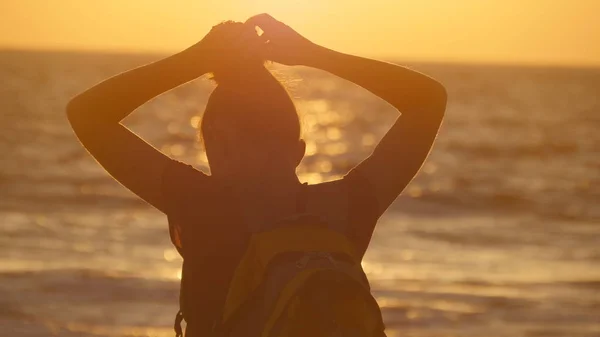 Jeune touriste attachant queue de cheval sur la plage près de la mer au coucher du soleil. Belle fille serrer ses cheveux au bord de l'océan au lever du soleil. Sac à dos femme tressant les cheveux et faisant une coiffure. Soins capillaires Vue arrière — Photo