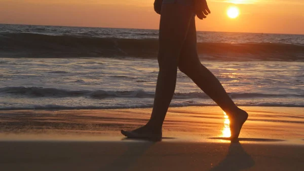 Ben av ung kvinna går längs ocean beach under sunrise. Kvinnliga fötter gå barfota på stranden vid solnedgången. Flicka kliva i grunt vatten vid strandlinjen. Sommar semester koncept. Närbild — Stockfoto