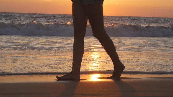 Pernas de mulher jovem indo ao longo da praia do oceano durante o nascer do sol. Pés femininos andando descalços na costa do mar ao pôr do sol. Rapariga a pisar em águas rasas na costa. Conceito de férias de verão. Fechar — Fotografia de Stock