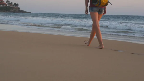 Piernas de viajera joven con mochila que va a lo largo de la playa del océano durante el amanecer. Pies de turista mujer caminando descalza en la orilla del mar al atardecer. Chica excursionista pisando durante los viajes o vacaciones de verano —  Fotos de Stock