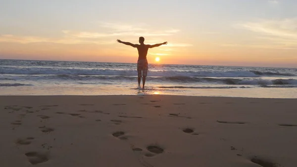 Giovane uomo che corre sulla spiaggia verso l'oceano al tramonto e alzò le mani. Ragazzo sportivo che va sulla riva sabbiosa al mare durante le vacanze. Rilassati durante le vacanze estive. Vista posteriore posteriore — Foto Stock