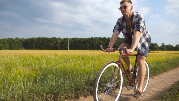 Sportlicher Typ mit Sonnenbrille radelt auf Feldwegen im Freien. Junger lächelnder Mann mit Oldtimer-Fahrrad auf Landstraße über Feld. Fahrradfahrer auf dem Land. Gesunder aktiver Lebensstil — Stockfoto