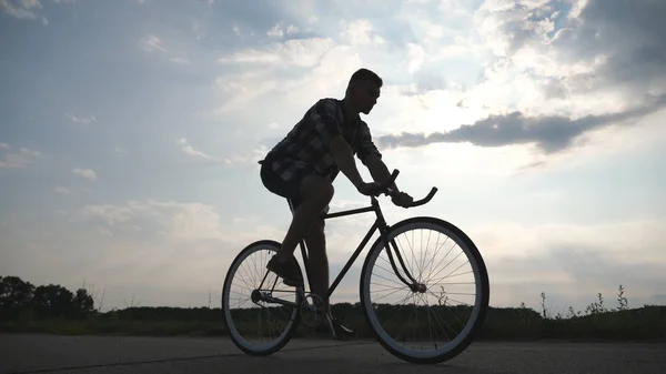 Silueta de hombre joven montando en bicicleta vintage con hermoso cielo al atardecer en el fondo. Tipo deportivo en bicicleta en la carretera del campo. Ciclista masculino montando bicicleta de engranaje fijo. Vida activa saludable Primeros planos — Foto de Stock