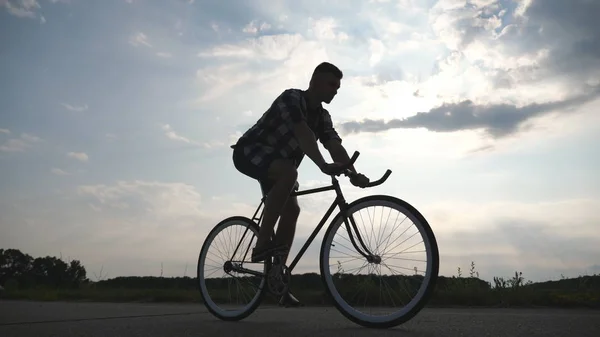 Silhouette eines jungen Mannes auf einem Oldtimer-Fahrrad mit wunderschönem Sonnenuntergangshimmel im Hintergrund. Sportlicher Typ beim Radeln auf der Landstraße. Radler mit festem Gang. gesunde aktive Lebensweise Nahaufnahme — Stockfoto