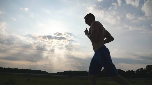 Silhouette eines muskulösen Mannes, der bei Sonnenuntergang auf der Landstraße joggt. Profil des männlichen Joggertrainings für Marathonläufe im Freien. Sportler, die vor blauem Himmel trainieren und laufen. Sport und aktiver Lebensstil — Stockfoto