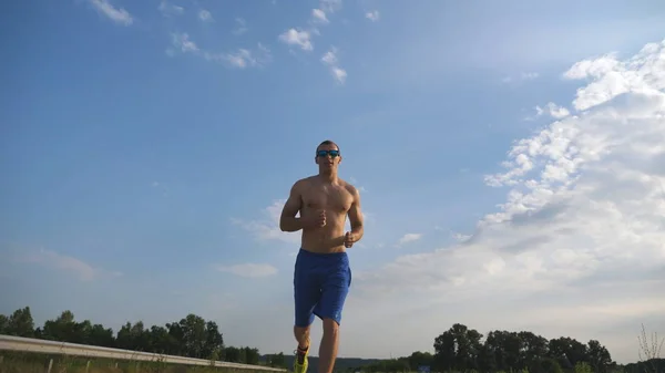 Homme musclé courant sur la route de campagne. Jeune athlète jogging à l'autoroute. Entraînement de sportif masculin sur la piste. Runner exercice en plein air. Mode de vie sain et actif Fermer — Photo