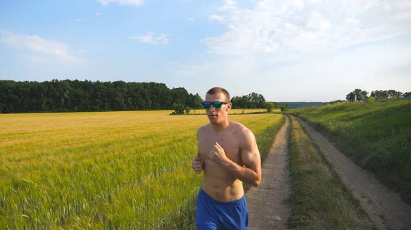 Muskulöser Mann, der auf Landstraße läuft. Junger, athletischer Kerl joggt auf ländlichem Pfad über das Feld. männliche Sportler beim Training in der Natur. Läufer, die im Freien trainieren. gesunder aktiver Lebensstil hautnah — Stockfoto