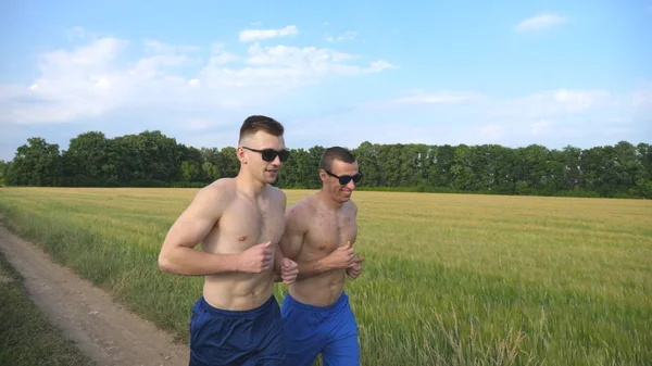 Dois homens musculados a correr e a falar ao ar livre. Jovens atletas sorridentes a correr pelo campo. Esportistas do sexo masculino treinando juntos na natureza. Amigos se exercitando lá fora. Estilo de vida ativo saudável — Fotografia de Stock