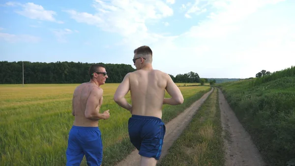 Två muskulös mannen köra och prata utomhus. Unga athletic killar jogging över fältet. Manliga sportsmans utbildning tillsammans på naturen. Vänner utövar. Hälsosam aktiv livsstil tillbaka bakifrån — Stockfoto