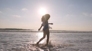 Okyanusun plajında yürürken mutlu kadın ve ayakları ile su püskürtme. Genç güzel kız yaşam zevk ve deniz kıyısında eğlenmek. Yaz tatili. Arka planda peyzaj