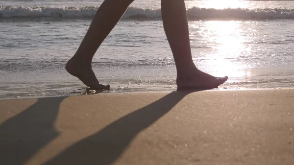 Nahaufnahme weiblicher Füße, die auf goldenem Sand am Strand mit Meereswellen im Hintergrund spazieren. Beine einer jungen Frau, die auf Sand tritt. Barfuß Mädchen am Ufer des Meeres. Sommerferienkonzept — Stockfoto