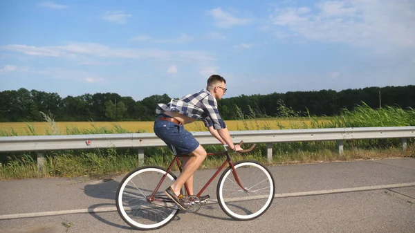 Joven hombre guapo montando en bicicleta vintage en la carretera del campo. Un tipo deportivo en bicicleta en la pista. Hombre ciclista montar bicicleta de engranaje fijo en la carretera. Vida activa saludable Movimiento lento — Foto de Stock