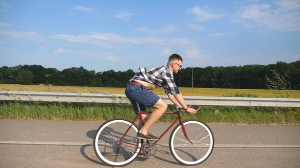 Joven hombre guapo montando en bicicleta vintage en la carretera del campo. Un tipo deportivo en bicicleta en la pista. Hombre ciclista montar bicicleta de engranaje fijo en la carretera. Vida activa saludable Movimiento lento — Foto de Stock