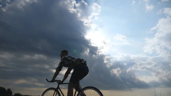 Silueta de hombre joven montando en bicicleta vintage con hermoso cielo al atardecer en el fondo. Tipo deportivo en bicicleta en la carretera del campo. Ciclista masculino montando bicicleta. Vida activa saludable Movimiento lento — Foto de Stock