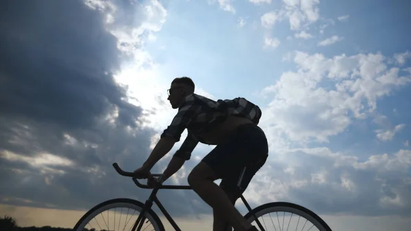 Silueta de hombre joven montando en bicicleta vintage con hermoso cielo al atardecer en el fondo. Tipo deportivo en bicicleta en la carretera del campo. Ciclista masculino montando bicicleta. Vida activa saludable Movimiento lento — Foto de Stock