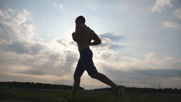 Silhouette of muscular man jogging in the country road at sunset. Male jogger training for marathon run outdoor. Athlete exercising and running against blue sky. Sport and active lifestyle Slow motion