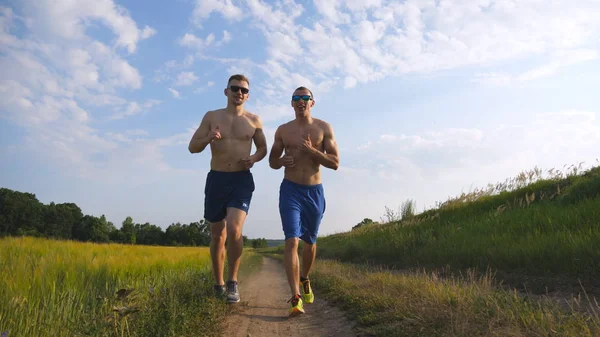 Two muscular men running and talking outdoors. Young athletic guys jogging over the field. Male sportsmans training together at nature. Friends exercising outside. Slow motion Healthy active lifestyle