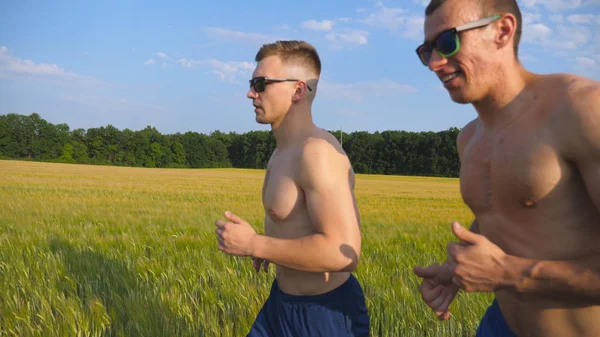 Dois homens musculados a correr ao ar livre. Jovens atletas a correr pelo campo. Esportistas do sexo masculino treinando juntos na natureza. Amigos se exercitando lá fora. Movimento lento Estilo de vida ativo saudável — Fotografia de Stock