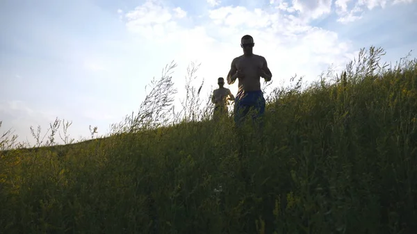 Groep jonge mannen lopen de groene heuvel over blauwe hemel met zon flare op de achtergrond. Mannelijke atleten is joggen in de natuur bij zonsondergang. Sport lopers gaan bergaf buiten bij zonsopgang. Slow Motion — Stockfoto