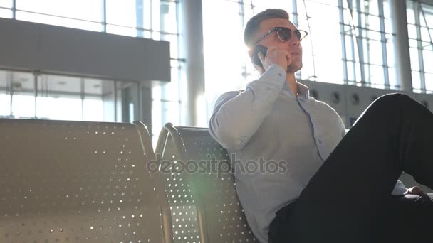 Joven hombre de negocios hablando por teléfono en la moderna terminal del aeropuerto con destello de sol en el fondo. Primer plano retrato de hombre de negocios guapo en gafas de sol hablando por teléfono celular en la oficina. Movimiento lento — Vídeo de stock