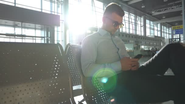 Молодой бизнесмен сидит в аэропорту и использует смартфон с солнечной вспышкой на заднем плане. Закрыть портрет красивого бизнесмена в солнцезащитных очках со смартфоном у терминала. Медленное движение — стоковое видео