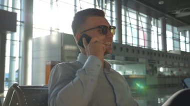 Havaalanında gülümseyen genç bir işadamı telefonda konuşuyor ve arka planda güneş parlaması var. Ofiste cep telefonuyla konuşan güneş gözlüklü mutlu iş adamının portresi. Yavaş çekim
