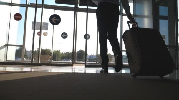 Uomo d'affari a piedi dall'aeroporto con i suoi bagagli. Uomo che passa attraverso la porta automatica di vetro per la strada e rotolare valigia su ruote. Il bagliore del sole sullo sfondo. Viaggio o concetto di viaggio. Rallentatore — Video Stock