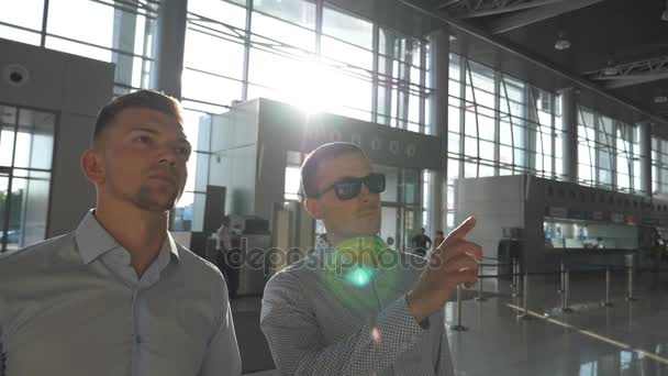 Uomo d'affari con gli occhiali da sole che indica qualcosa al suo collega. Due giovani uomini d'affari che guardano gli orari dei voli all'aeroporto. Luminoso sole che splende attraverso la finestra sullo sfondo. Rallentatore Close up — Video Stock