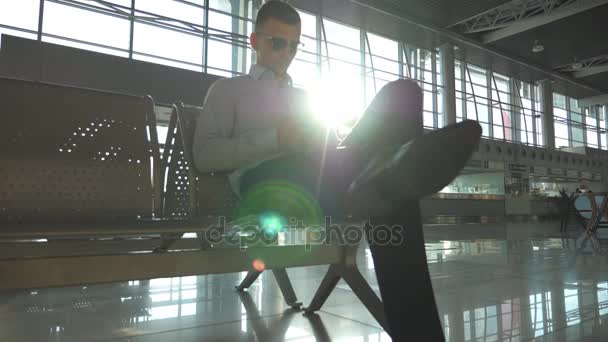 Giovane uomo d'affari seduto in aeroporto e utilizzando smartphone con brillamento solare sullo sfondo. Ritratto ravvicinato di un bell'uomo d'affari con occhiali da sole e smartphone al terminal. Rallentatore — Video Stock