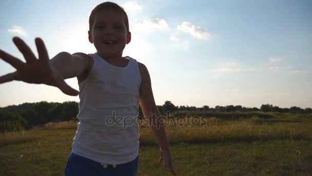 Menino feliz com uma mão estendida correndo no campo atrás da câmera e tentando pegá-lo. Sorrindo garoto do sexo masculino se divertindo na natureza no dia ensolarado. Criança a correr ao ar livre. Lento movimento Fechar — Vídeo de Stock