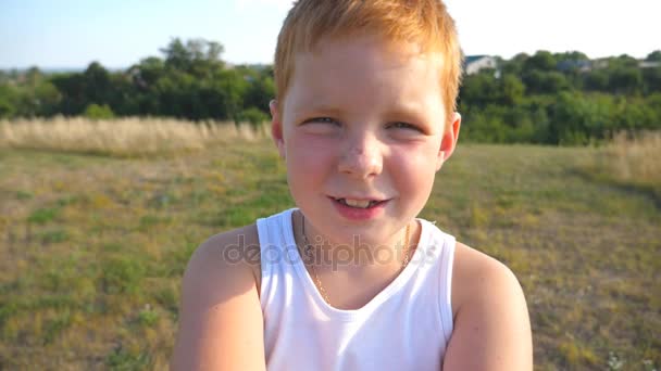 Gros plan sur les émotions de l'enfant mâle avec une expression heureuse sur le visage. Portrait de garçon aux cheveux roux heureux avec des taches de rousseur en plein air. Adorable beau bébé regardant dans la caméra avec un sourire joyeux. Mouvement lent — Video