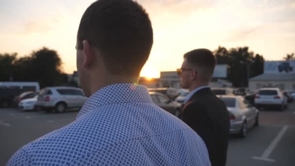 Geschäftsleute, die bei Sonnenaufgang zur Arbeit pendeln. selbstbewusste Typen, die auf dem Weg ins Amt sind. zwei junge Geschäftsleute zu Fuß auf einem Parkplatz mit Sonne im Hintergrund. Kollegen gehen nach draußen. Zeitlupe — Stockvideo