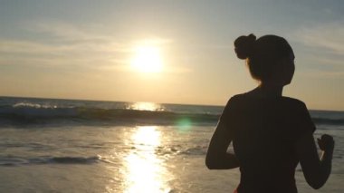 Güneş doğarken okyanus kıyısında koşan bir kız. Gün batımında sahilde koşan genç bir kadının silueti. Dişi sporcu dışarıda egzersiz yapıyor. Doğada sağlıklı, aktif bir yaşam tarzı. Yavaş çekim