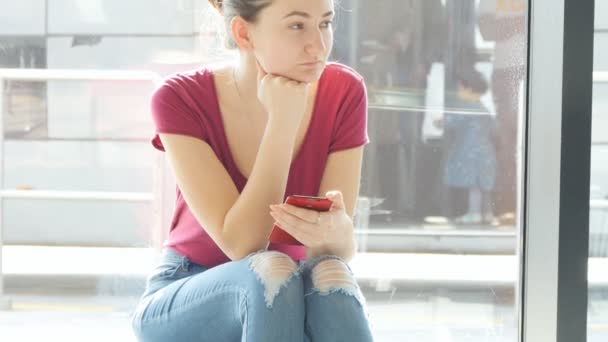 Jeune femme assise près de la fenêtre au salon d'attente de l'aéroport et regardant autour, puis en utilisant un smartphone. Attentes de vol à l'aéroport. Gros plan — Video