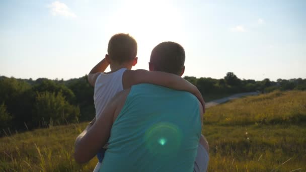 Der Vater verbringt mit seinem kleinen Sohn die Zeit im Freien bei Sonnenuntergang. Vater und Kind sitzen im grünen Gras auf dem Hügel und blicken auf die Natur um sie herum. Zeitlupe Rückansicht — Stockvideo