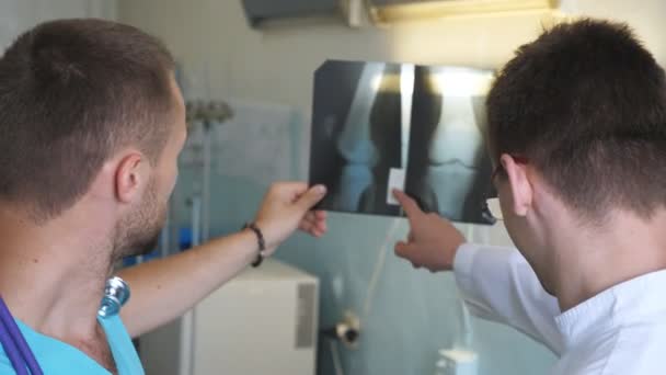 Два врача-мужчины рассматривают снимок МРТ и обсуждают его. Медработники в больнице ищут и анализируют рентгеновские снимки. Медики консультируются друг с другом. Закрыть задний вид — стоковое видео