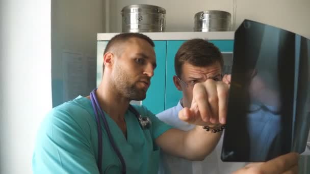 Médecins masculins se consultent tout en regardant l'image de rayons X. Deux médecins caucasiens regardent la photo IRM et en discutent. Les travailleurs médicaux à l'hôpital examinent les empreintes radiographiques. Gros plan — Video