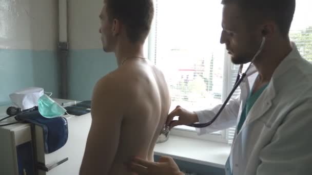 Medic controleren van rug en borst van de man in haar kantoor in het ziekenhuis. Examencommissie jonge mannelijke patiënt arts met de stethoscoop. Medisch-werker luisteren heartbeat van atletische man. Close-up — Stockvideo