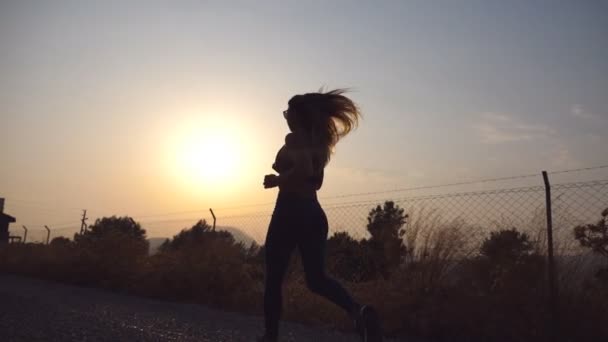 Seguindo para jogging menina desportiva na estrada do campo ao nascer do sol. Jovem a correr ao ar livre de manhã. Estilo de vida ativo saudável. Movimento lento — Vídeo de Stock