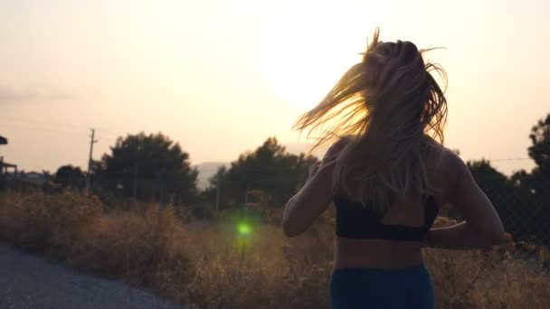 Siga a la chica deportiva trotando en la carretera del campo. Mujer joven en gafas de sol corriendo al aire libre y escuchando música desde el teléfono inteligente. Vida activa saludable. Vista trasera trasera en cámara lenta — Vídeos de Stock