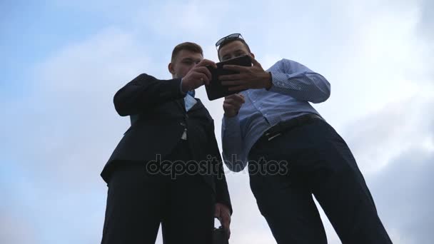 Deux jeunes hommes d'affaires parlant et utilisant tablette PC en plein air. Les hommes d'affaires travaillant sur tablette numérique à l'extérieur avec ciel en arrière-plan. Chers collègues appliquant la technologie mobile. Angle de vue bas Mouvement lent — Video
